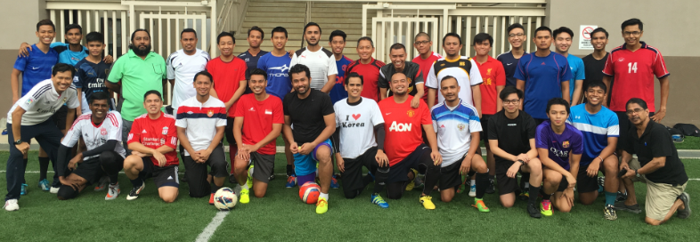 A large group of men in colorful soccer jerseys posing together on an indoor turf field with a soccer ball.
